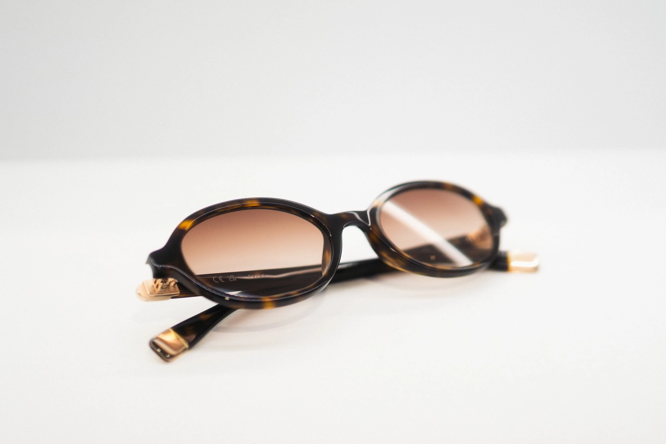 Occhiali da sole ovali con montatura tartarugata scura, lenti marroni sfumate e dettagli dorati sulle astine.