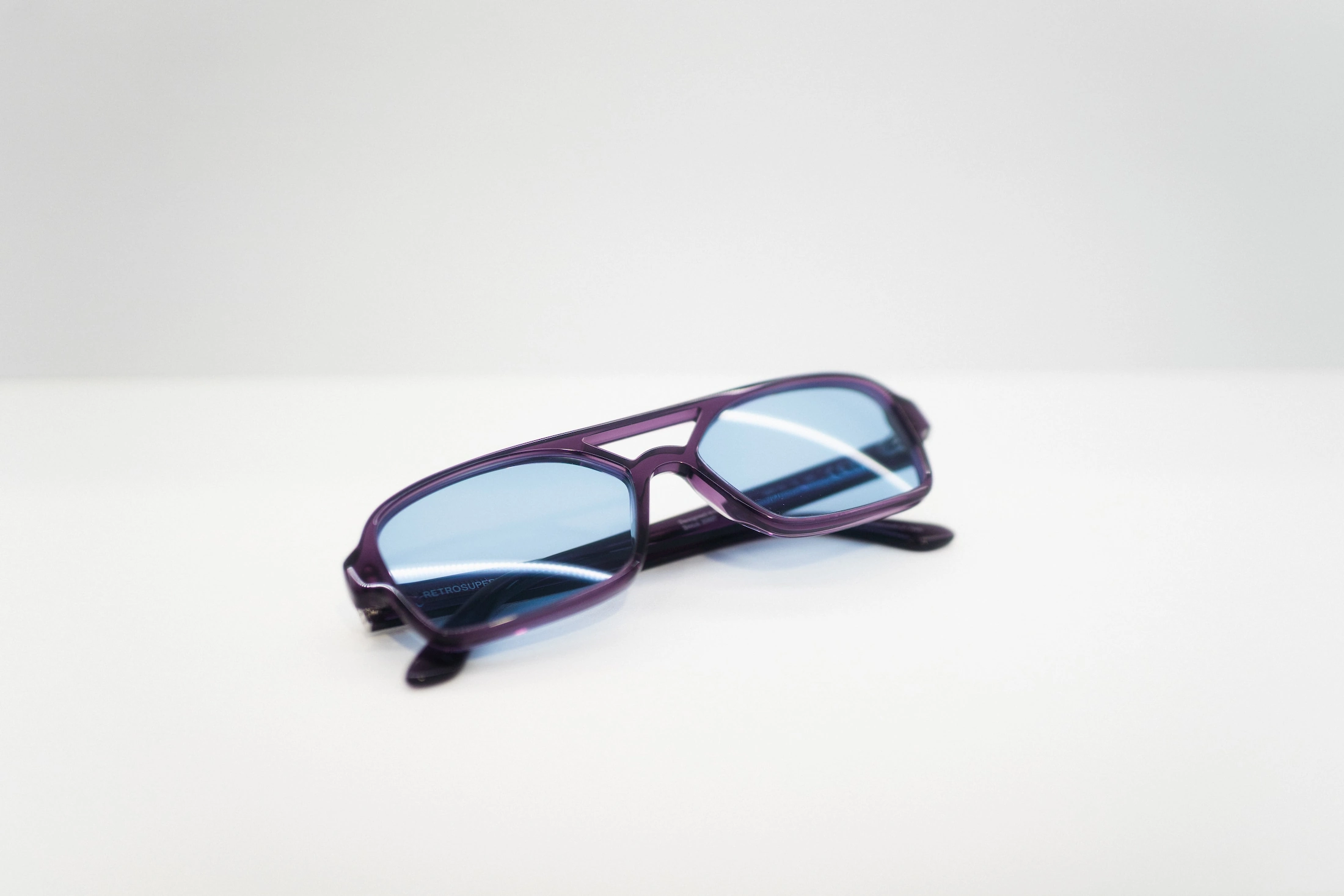 Occhiali da sole geometrici con doppio ponte, montatura viola trasparente e lenti azzurre chiare.