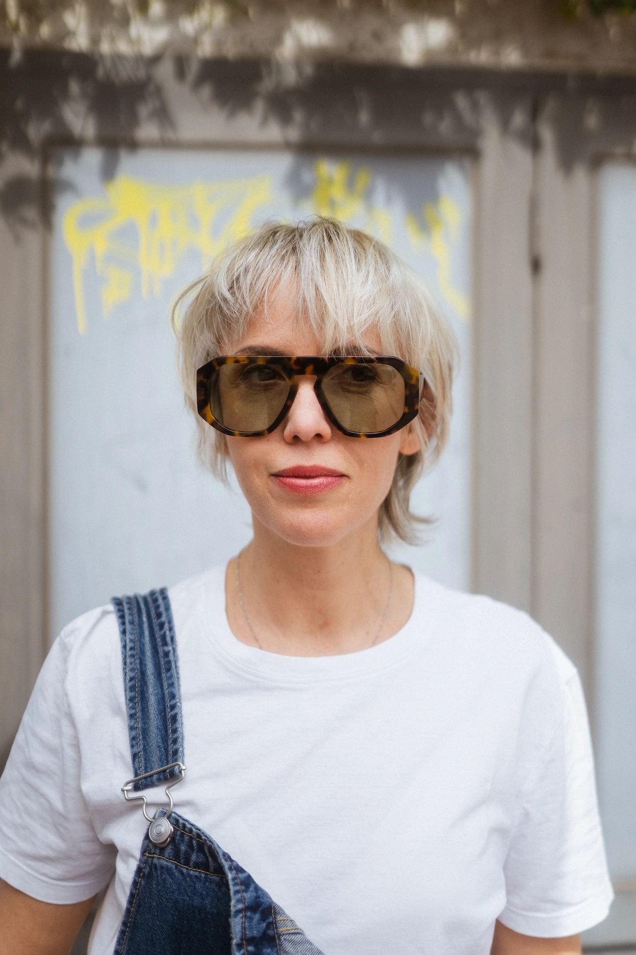 Donna in stile edgy con giacca argentata metallizzata e tatuaggi, indossa occhiali da sole neri cat-eye sotto una luce solare diretta con ombre di foglie sullo sfondo.