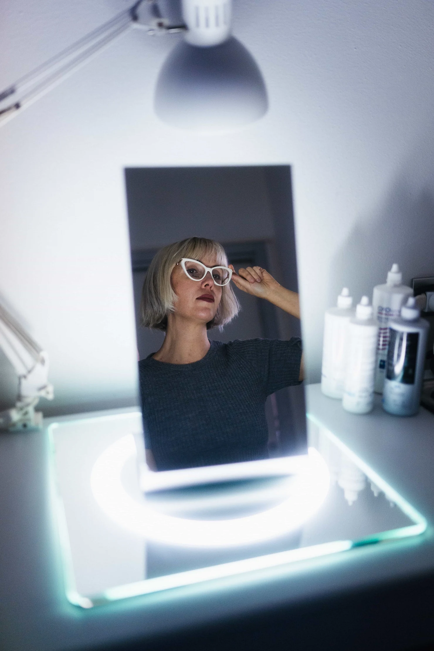 Riflesso nello specchio di una donna con caschetto biondo che prova una montatura di occhiali bianca stile cat-eye in un ambiente luminoso.