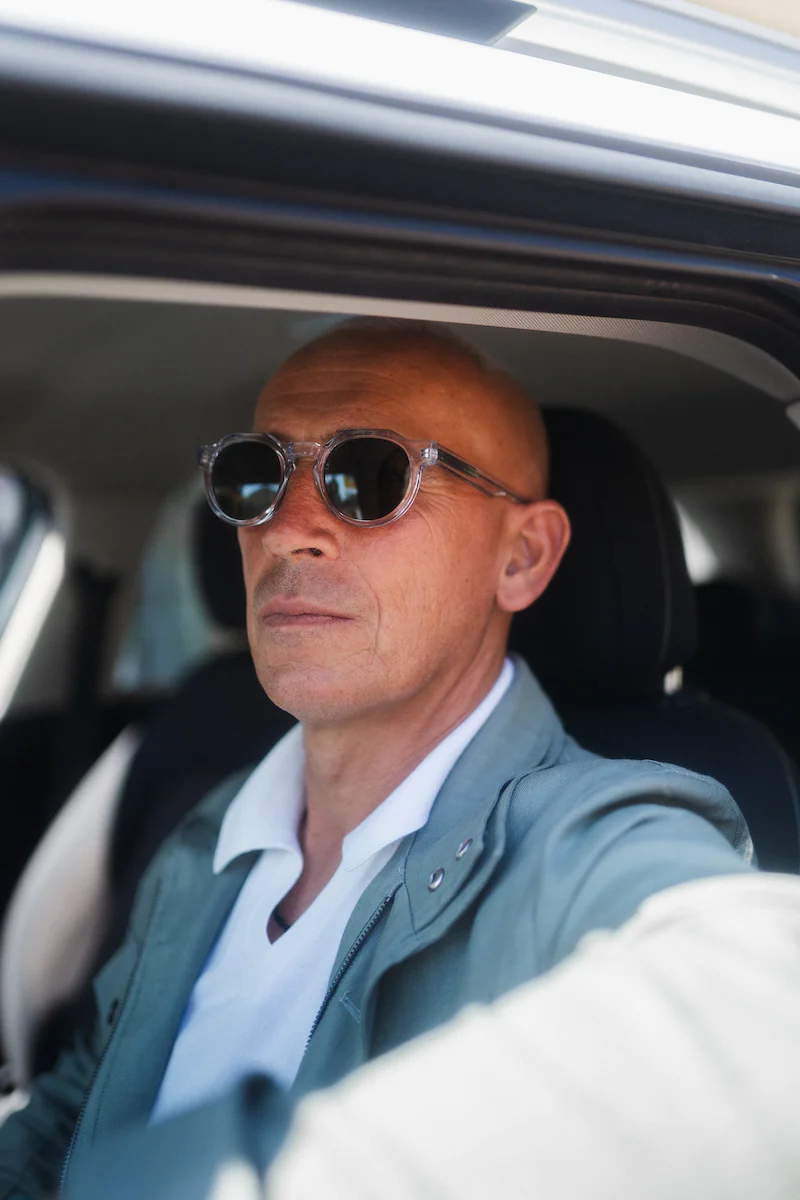 Uomo alla guida di un'auto con occhiali da sole dalla montatura trasparente, illuminato dalla luce che entra dal tetto apribile.