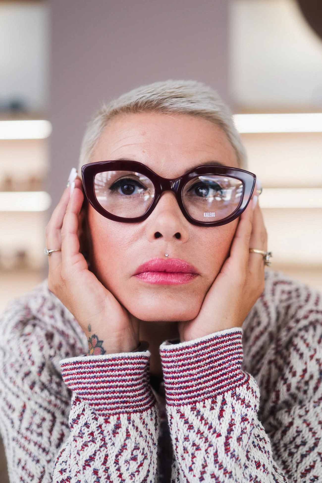 Primo piano di donna con capelli corti biondi che indossa occhiali da vista spessi color bordeaux, con le mani sul viso e un maglione fantasia.