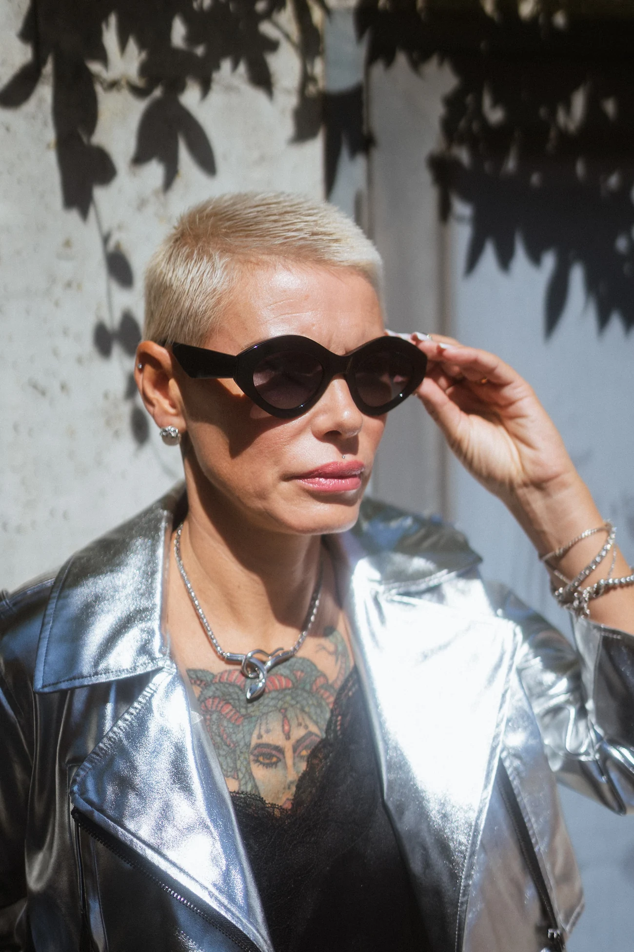 Donna in stile edgy con giacca argentata metallizzata e tatuaggi, indossa occhiali da sole neri cat-eye sotto una luce solare diretta con ombre di foglie sullo sfondo.