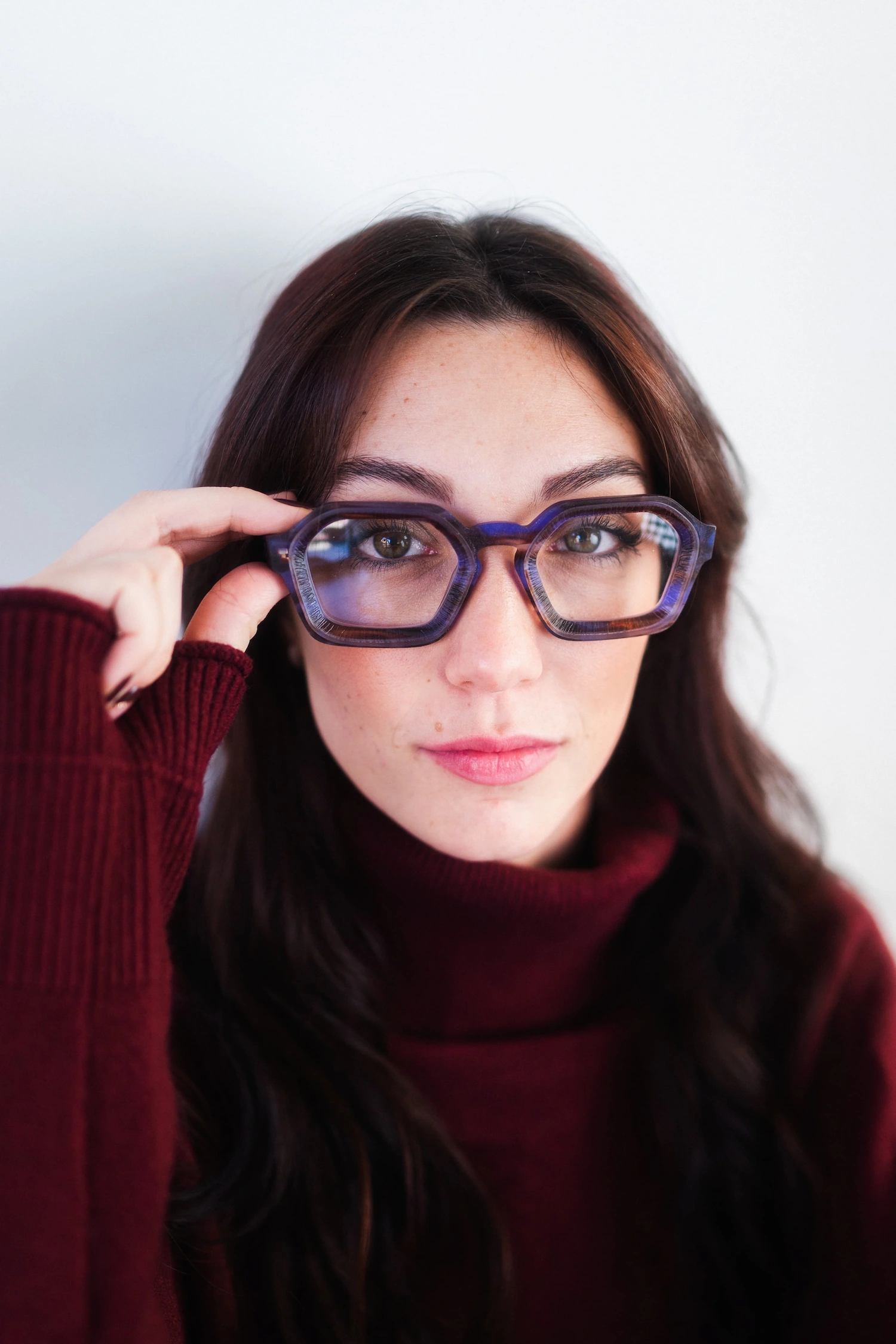 Ritratto ravvicinato di ragazza con maglione collo alto bordeaux che indossa occhiali da vista geometrici esagonali con montatura viola e blu.