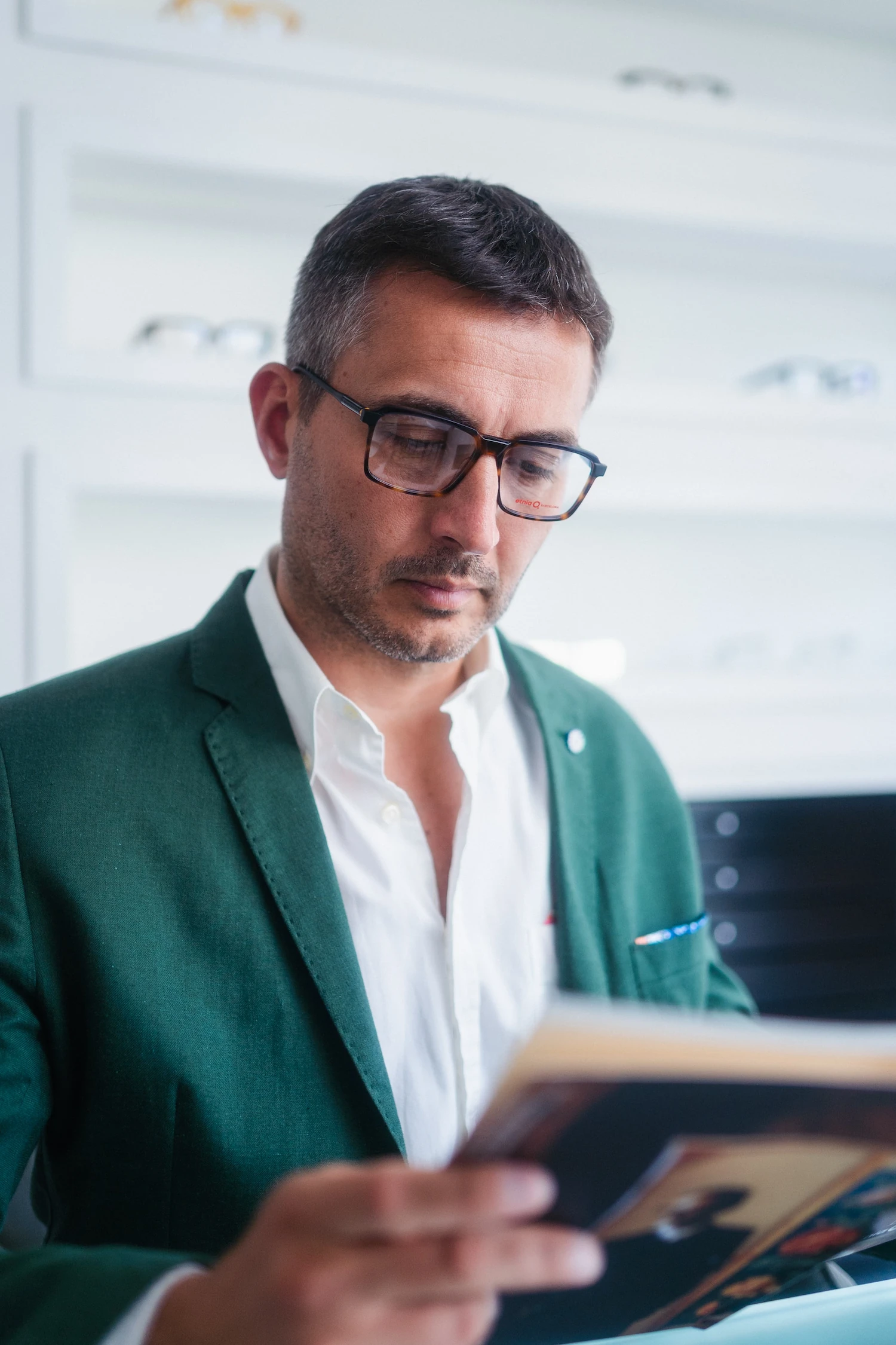 Uomo in giacca verde smeraldo che legge una rivista all'interno di un negozio di ottica, indossando occhiali da vista rettangolari tartarugati.