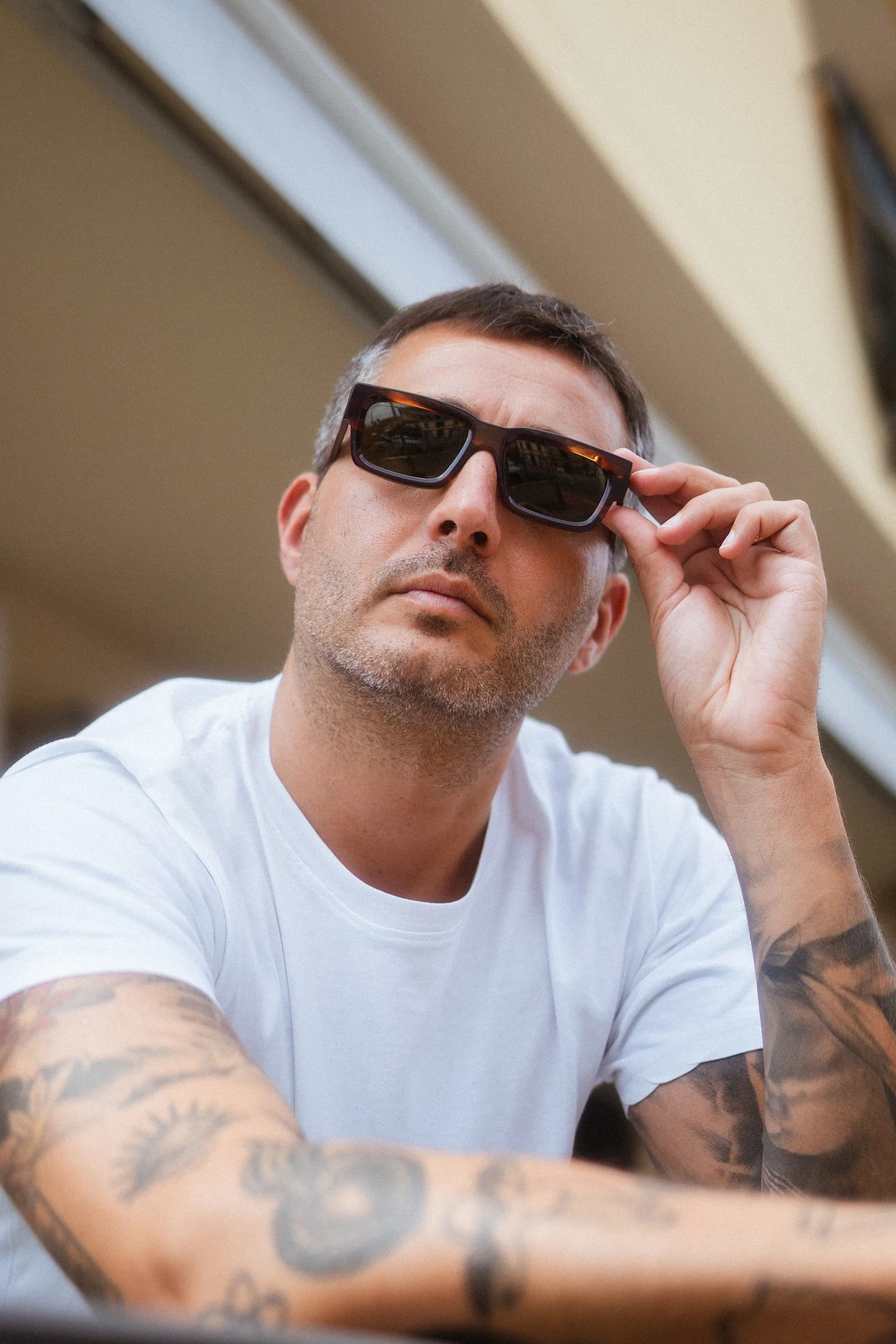 Ritratto lifestyle all'aperto di un uomo con braccia tatuate e t-shirt bianca che si aggiusta gli occhiali da sole rettangolari scuri.