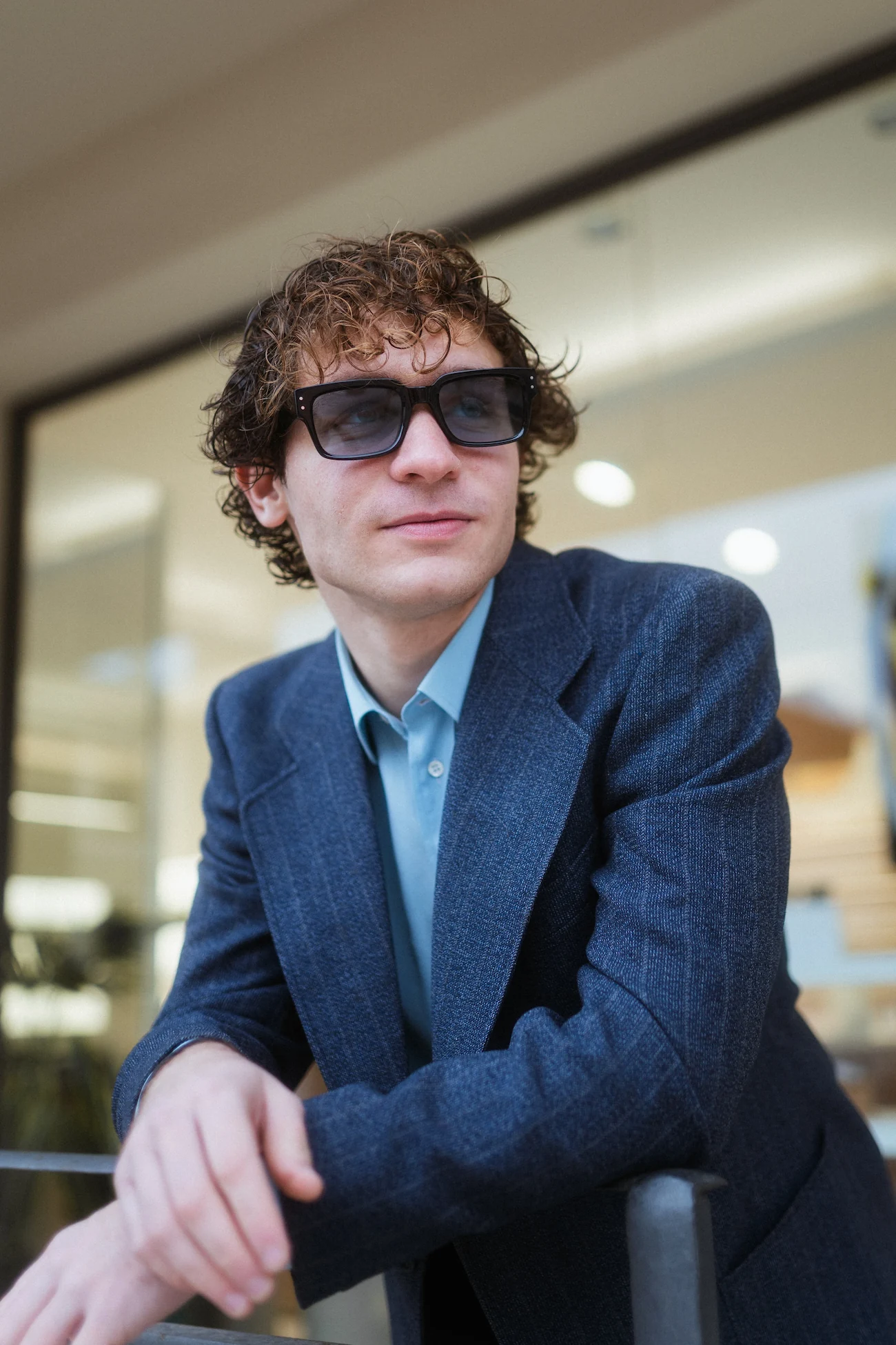 Giovane uomo con capelli ricci e giacca blu elegante in posa all'esterno, indossa occhiali da sole scuri con lenti leggermente azzurrate.