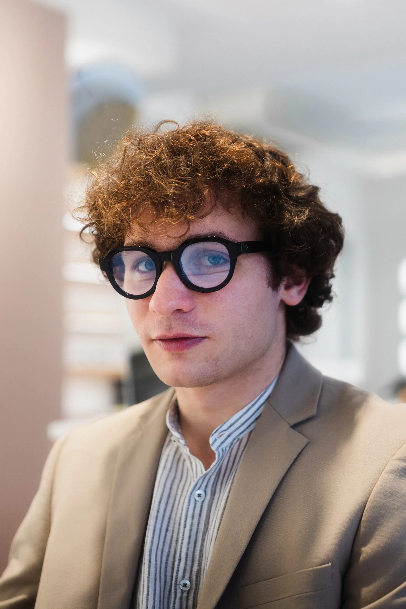 Ritratto in interno di giovane professionista con giacca beige e camicia a righe che indossa spessi occhiali da vista neri rotondi.