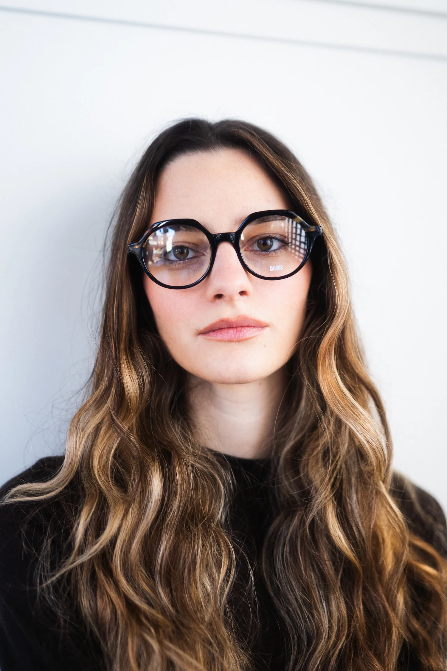 Foto ritratto frontale minimalista di ragazza con lunghi capelli mossi che indossa occhiali da vista neri dalla forma arrotondata su sfondo bianco.