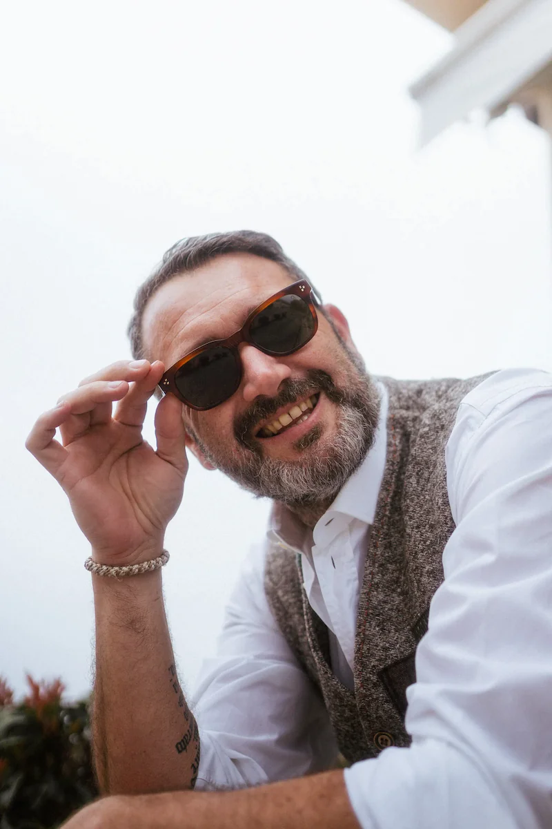 Uomo sorridente in stile classico con gilet in tweed e camicia bianca, tocca la montatura dei suoi occhiali da sole marroni guardando in camera.