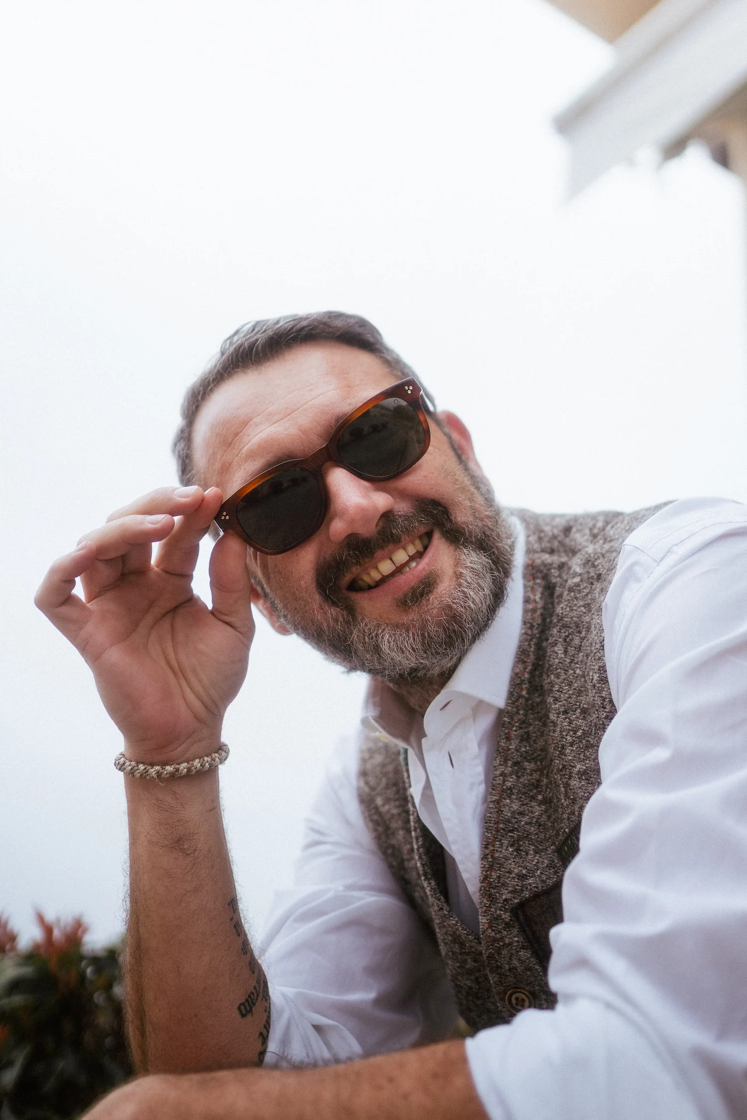 Uomo sorridente in stile classico con gilet in tweed e camicia bianca, tocca la montatura dei suoi occhiali da sole marroni guardando in camera.